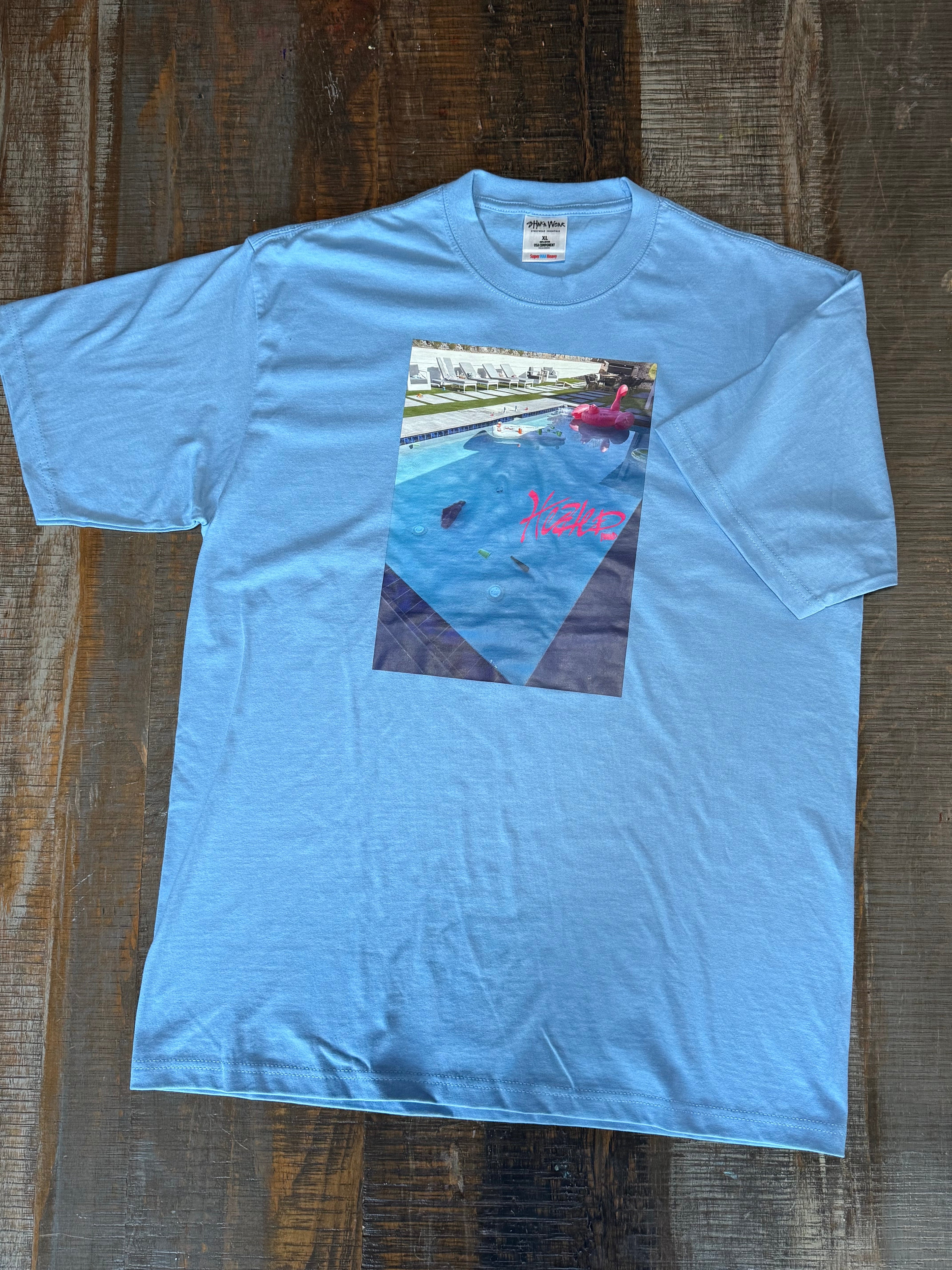 Light blue t-shirt with a graphic of a pool and text on a wooden surface