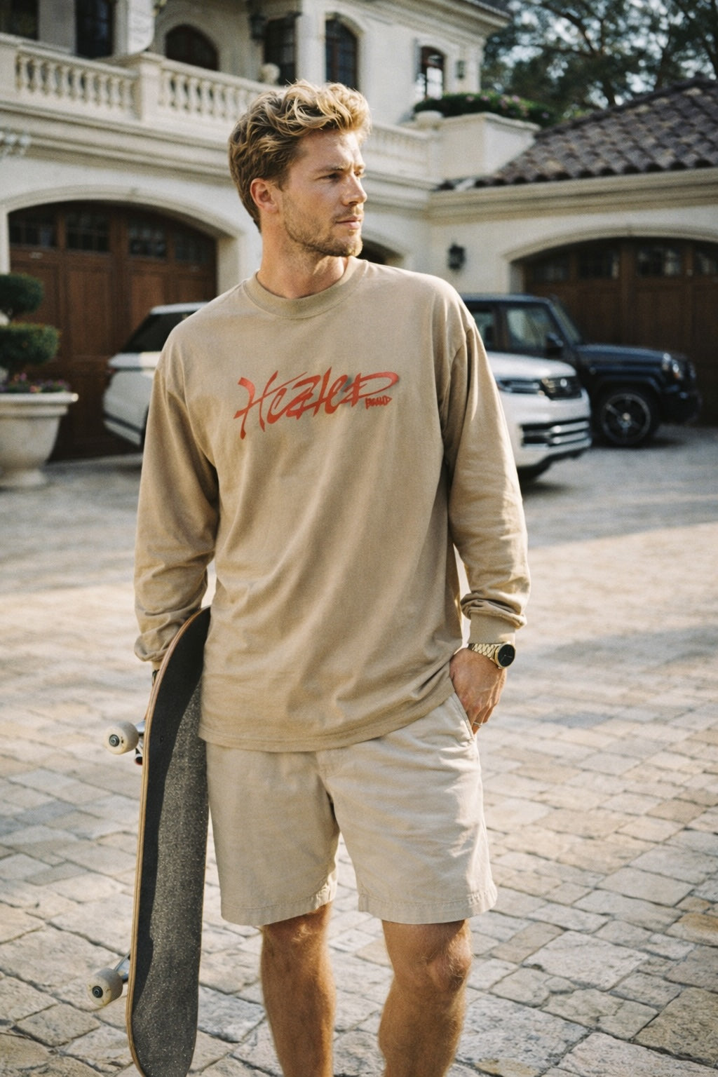 Man wearing a beige sweatshirt with red text, holding a skateboard, standing in a driveway.