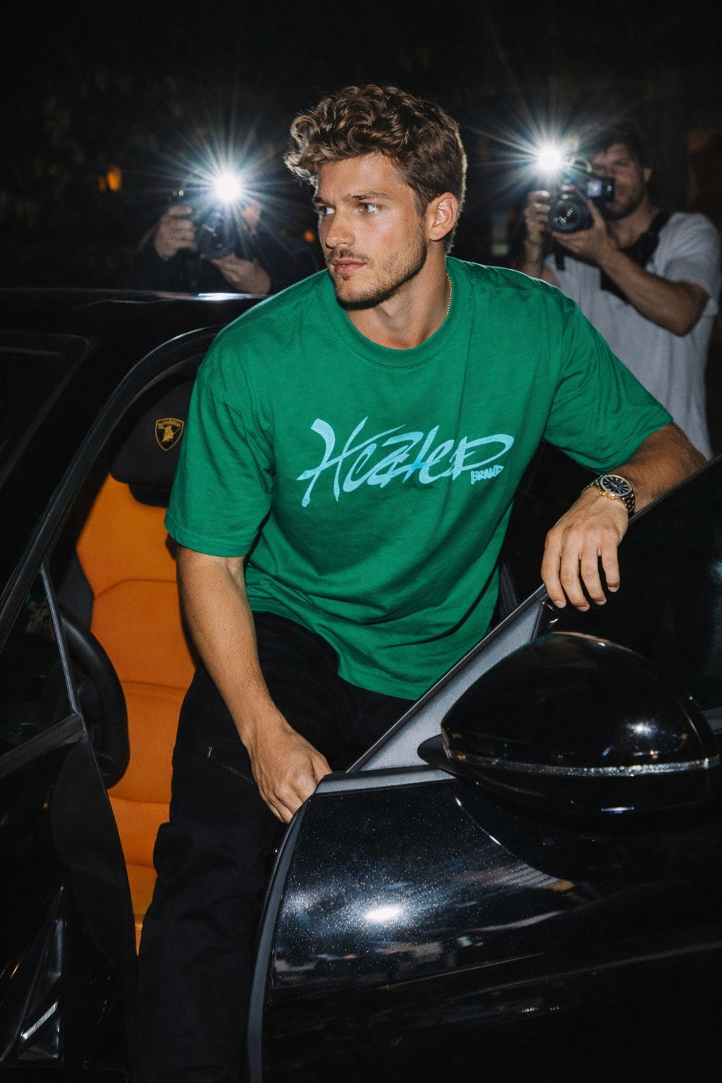 Man in a green t-shirt with 'Heated' logo standing next to a lambo at night.