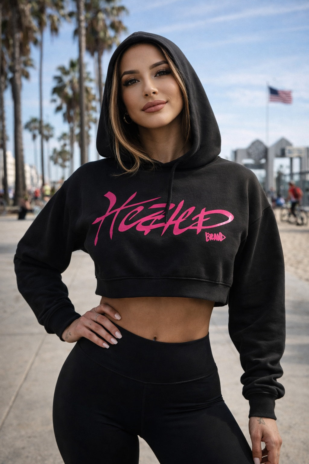 Woman wearing a black hoodie with pink text in an urban setting with palm trees and buildings.