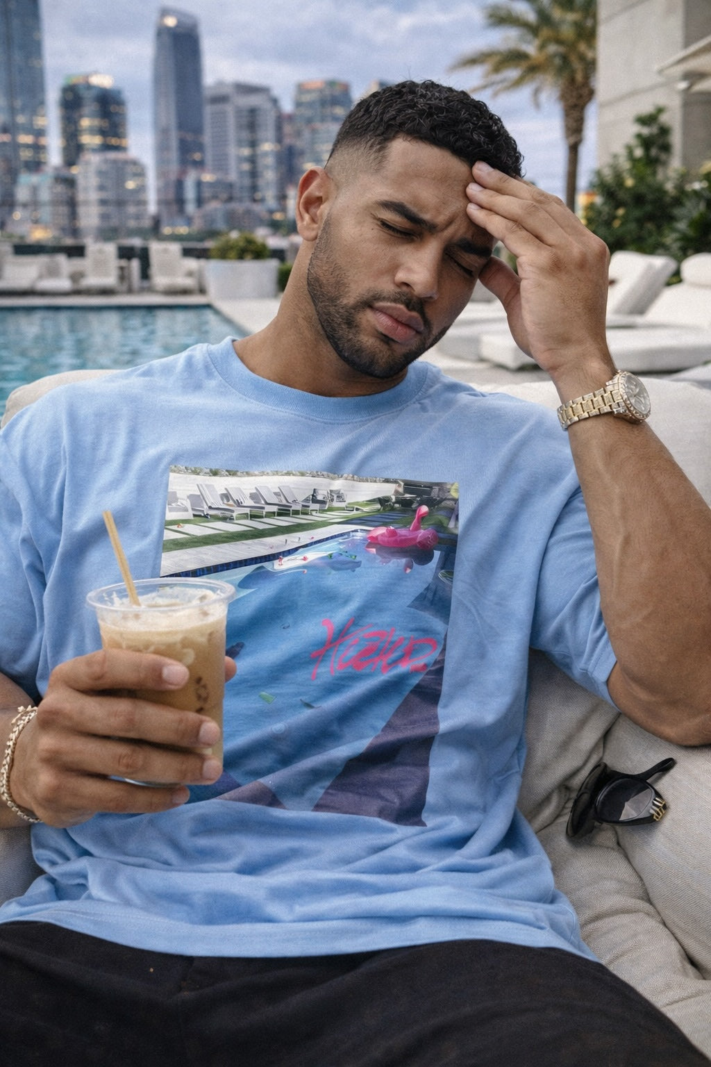 Man in a blue t-shirt holding a drink by a pool with cityscape in the background